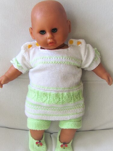 Babypuppe trägt ein gestricktes weiß-hellgrünes Set mit Oberteil, kurzer Hose und Schühchen mit Marienkäfern.
