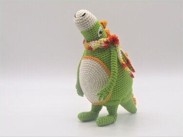 Gehäkelter grüner Drache mit cremefarbenem Bauch und mehrfarbigem Schal auf neutralem Hintergrund.