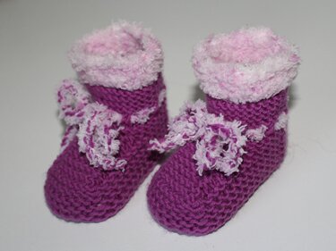 Zwei lila gestrickte Babyschuhe mit rosa-weißem Plüschrand und Schleifen auf hellem Hintergrund
