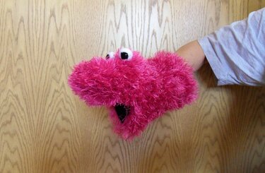 Pinke flauschige Handpuppe mit großen Augen und offenem Maul, auf einer Hand getragen vor hellem Holz.