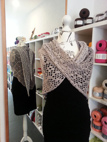 Hellbeiges gehäkeltes Dreieckstuch mit offenem Lochmuster, drapiert auf einer Schaufensterpuppe; Garnregal und Spiegelung im Hintergrund