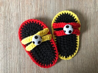 Zwei gehäkelte Baby-Fußballschühchen in Schwarz, Rot und Gelb mit Fußballknöpfen.