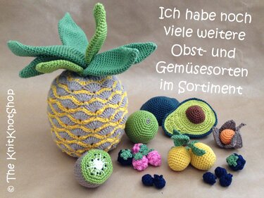 Gehäkelte Ananas, aufgeschnittene Avocado, Kiwi, Zitronen, Him- und Heidelbeeren auf beigem Untergrund.