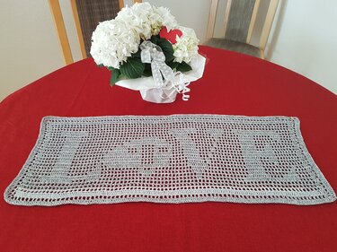 Gehäkelter rechteckiger Filet-Tischläufer mit Buchstabenmotiv auf rotem Tischtuch und weißer Hortensie