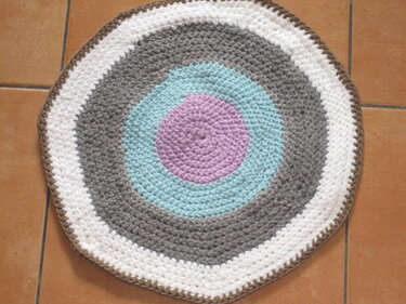 Runder, gehäkelter Teppich mit konzentrischen Ringen in Lila, Hellblau, Grau und Weiß auf gefliestem Boden.