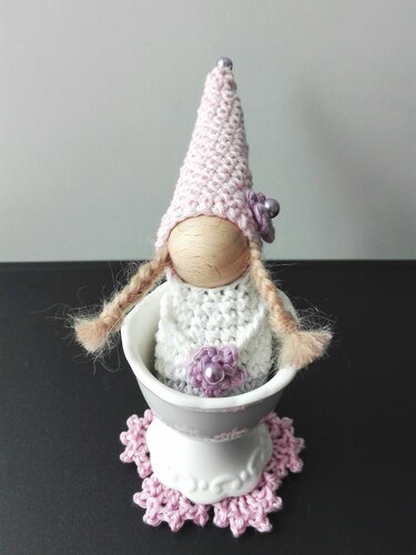Gehäkelter Amigurumi-Wichtel mit Holzgesicht, rosa spitzer Mütze und geflochtenen Zöpfen im weißen Eierbecher auf rosa Häkeluntersetzer.