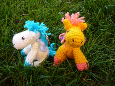 Zwei gehäkelte Pony‑Schlüsselanhänger (gelb und weiß) auf Gras mit bunten Mähnen
