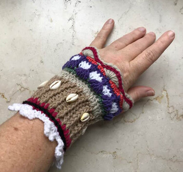 Hand mit bunt gestreifter, gehäkelter fingerloser Armstulpe und aufgenähten Kaurimuscheln