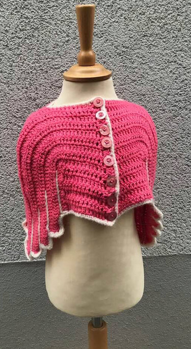 Kurz geschnittenes rosa gehäkeltes Kindercape mit sechs Knöpfen und weißem Zackenrand