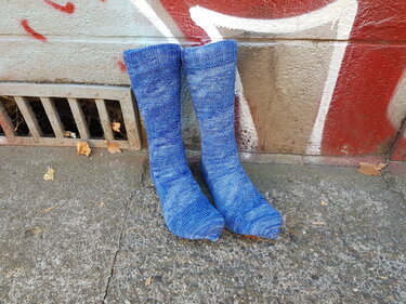 Paar hohe, handgestrickte blaue Herrensocken vor einer Wand mit Graffiti auf Betonboden.