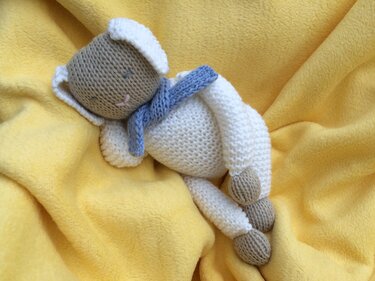 Gestricktes Amigurumi‑Schäfchen mit blauem Schal, schlafend auf gelber Decke