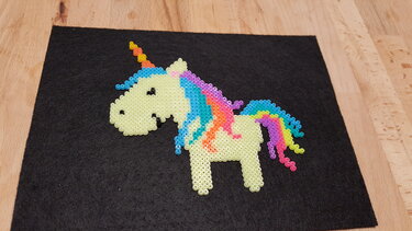 Einhorn aus Bügelperlen mit cremefarbenem Körper und bunter Regenbogenmähne auf schwarzem Filz