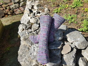 Mehrfarbige Stricksocken mit Strukturmuster auf einer Steinfläche vor Ziegeln und Vegetation.