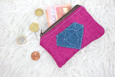 Nahaufnahme einer pinken Stofftasche mit braunem Reißverschluss und blau gestickter Diamant-Applikation, daneben Euro-Münzen und -Scheine auf weißem Untergrund