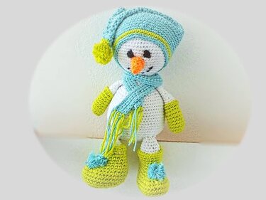 Gehäkelte Schneemann-Puppe in Weiß mit hellblauer Mütze, Schal sowie grünen Fäustlingen und Stiefeln.