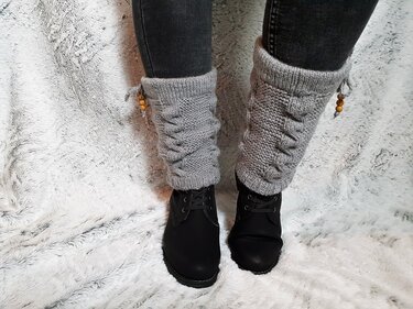 Beine in Jeans und schwarzen Stiefeln mit grauen gestrickten Beinstulpen, Zopfmuster und Kordel mit Holzperlen
