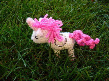 Gehäkeltes weißes Einhorn mit rosa Mähne und Metallring auf grünem Gras.