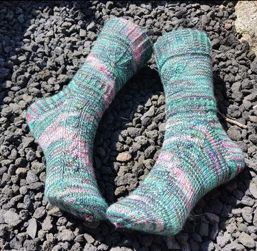 Paar handgestrickter Socken in Türkis, Rosa und Violett auf dunklem Kies.