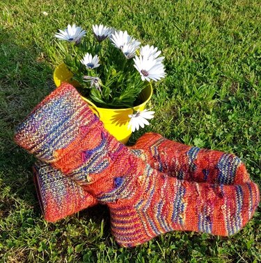 Mehrfarbige gestrickte Damensocken in Orange-Rot auf Gras neben gelbem Blumentopf mit weißen Blumen