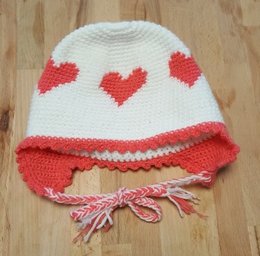 Weiße gehäkelte Babymütze mit korallenfarbenen Herzen, Ohrenklappen und geflochtenen Bindebändern auf Holzuntergrund