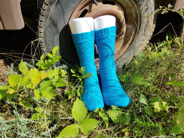 Türkisfarbene gestrickte Herrensocken auf weißen Sockenformen im Gras vor einer rostigen Felge