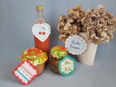 Gehäkelte Geschenkanhänger an Flasche und Gläsern mit Kirschmotiv sowie den Aufschriften „Frohe Ostern“ und „Alles Liebe“, daneben Trockenblumen
