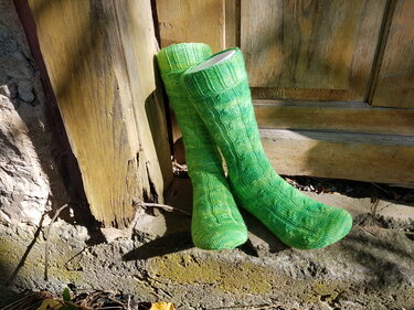 Zwei lange grüne, gestrickte Herrensocken mit Zopfmuster, an eine Holztür gelehnt