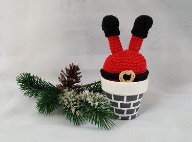 Gehäkelter Weihnachtsmann mit roten Beinen und schwarzen Stiefeln, der aus einem grau bemalten Schornstein-Topf ragt, daneben ein Tannenzweig und ein Zapfen auf weißem Hintergrund.