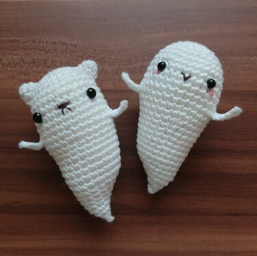 Zwei weiße gehäkelte Amigurumi-Geister, eines mit Katzenohren, auf Holzuntergrund.