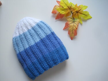 Gestrickte Babymütze in Weiß, Hellblau und Blau liegend auf hellem Untergrund neben künstlichen Herbstblättern