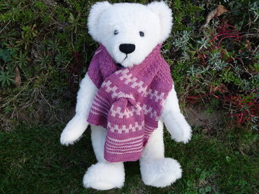 Weißer Teddybär mit gestrickter lila Mütze und passendem Schal im Zickzack-Muster auf grünem Untergrund.