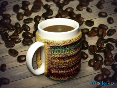 Weiße Kaffeetasse mit braun-grün gestreifter gehäkelter Hülle, umgeben von Kaffeebohnen auf Holztisch.