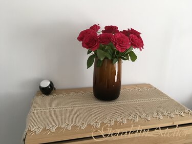 Beiger gehäkelter Tischläufer mit Netzmuster und gezackter Spitzenkante auf Holzkommode, braune Vase mit roten Rosen