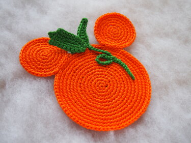 Bright orange crocheted pumpkin-shaped coaster with green stem and leaf on a white background.
