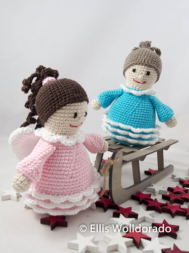 Zwei gehäkelte Amigurumi-Engelchen in Rosa und Hellblau neben einem Holzschlitten und verstreuten Holzsternen