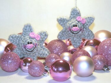 Zwei gehäkelte graue Sterne mit rosa Schleifen und lächelnden Gesichtern, umgeben von rosa Weihnachtskugeln.
