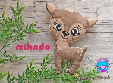 Braunes Stoff-Rehkitz mit großen Augen und weißen Punkten vor Holz und grünen Blättern.