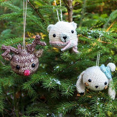 Drei gehäkelte Tierkopf-Anhänger an Tannenzweigen: Rentier, Bär mit Schal und Häschen mit blauer Mütze