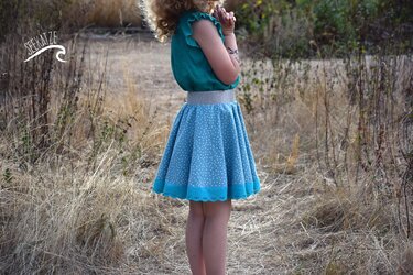 Seitliche Ansicht eines Kindes in hellblauem, weiß gepunktetem Tellerrock mit türkisfarbener Spitze und elastischem Glitzerbund