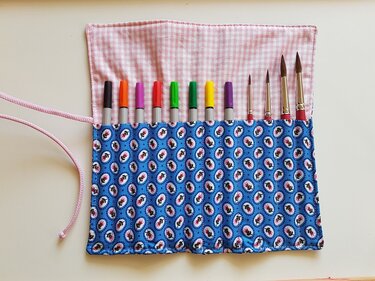 Geöffnete Stifterolle mit schmalen Fächern, Bindeband, blauem Außenstoff und rosa Innenfutter, bestückt mit Stiften und Pinseln