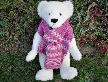 Weißer Plüschteddy mit pink-weiß gemustertem Strickschal und passender Mütze, liegend im Gras.