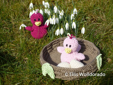 Zwei gehäkelte Amigurumi-Vögel: rosa im Nest mit Eiern, magenta im Gras bei Schneeglöckchen.