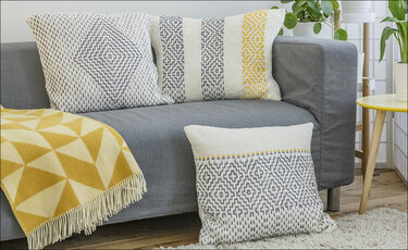 Drei gestrickte Kissen und eine Decke auf einem grauen Sofa in Weiß, Grau und Gelb mit geometrischen Mustern.