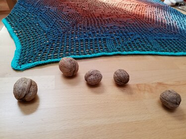 Gehäkeltes Tuch mit Farbverlauf von Türkis zu Rostrot und türkiser Kante auf hellem Holztisch mit Walnüssen.