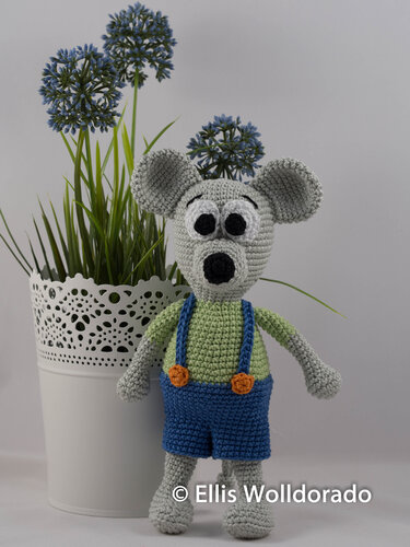Gehäkelte graue Amigurumi‑Maus mit grünem T‑Shirt und blauer Latzhose neben weißem Blumentopf