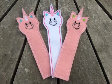 Drei Filz-Einhorn-Lesezeichen in Pastellfarben mit gestickten Konturen auf einem Holztisch