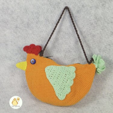 Gehäkelte orange Hühnertasche mit rotem Kamm, gelbem Schnabel, blauem Knopfauge, hellgrünen Flügeln, braunem Trageriemen und Reißverschluss