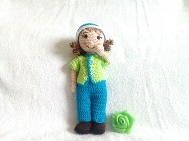 Amigurumi-Puppe mit grünem Cardigan, blauer Hose, braunen Schuhen, lockigem Haar und grüner Häkelrose, stehend auf weißem Untergrund.