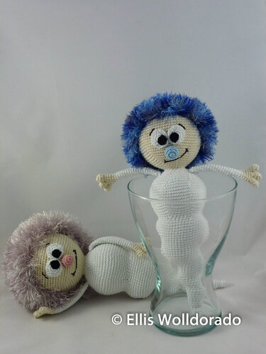 Zwei gehäkelte Amigurumi-Babys mit flauschigem Haar, eines in einem Glas, das andere liegend vor hellem Hintergrund