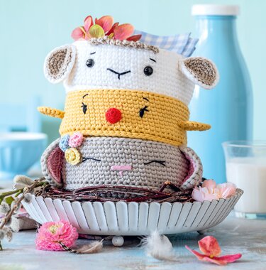 Drei gestapelte gehäkelte Tierkörbe im Amigurumi‑Stil mit Blumenapplikationen und Pastellfarben auf einer Schale